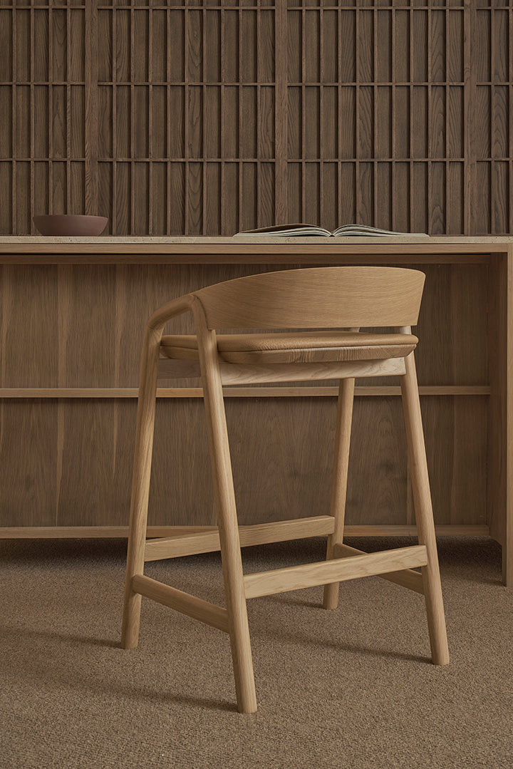 The Inlay Counter Stool by Wooden Stories, featuring a leather seat, is placed by a wooden desk with an open book and bowl, set against wood paneling and shelves in a warm, neutral-toned room.