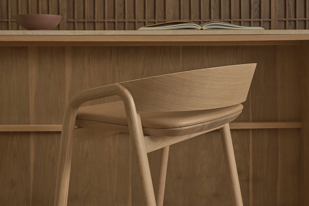 The Inlay Counter Stool with leather seat by Wooden Stories stands before a wooden desk. An open book and brown bowl are on the desk, while a wood panel wall forms a warm, inviting backdrop.