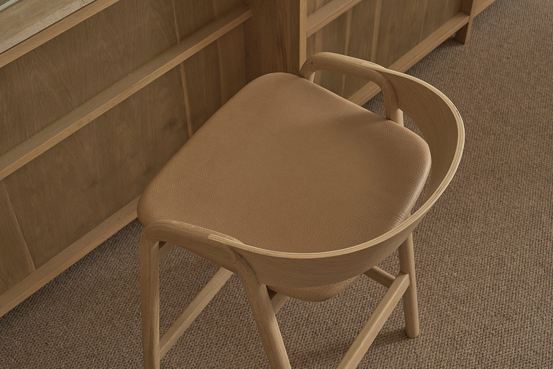 A modern Wooden Stories Inlay Counter Stool with a leather seat sits on a brown carpet, next to a matching solid oak desk.