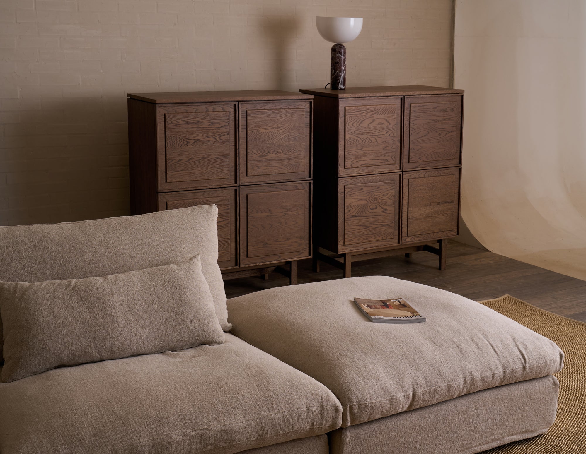 A cozy living room with a beige sectional sofa and a magazine on the ottoman. Behind the sofa are two Wooden Stories Yorke Highboards with square panels, and a modern lamp with a white globe shade sits atop one cabinet.