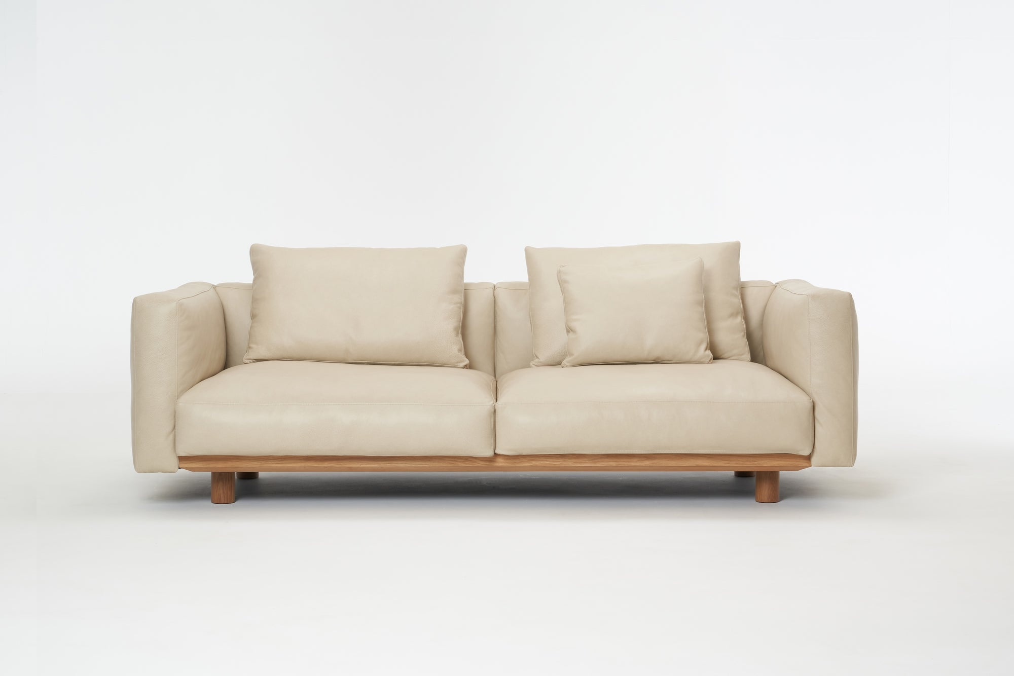 The Wooden Stories Islet Sofa | Leather is a modern beige sofa with durable leather, two seat cushions, two large backrest pillows, two smaller pillows, and a wooden base on a plain white background.