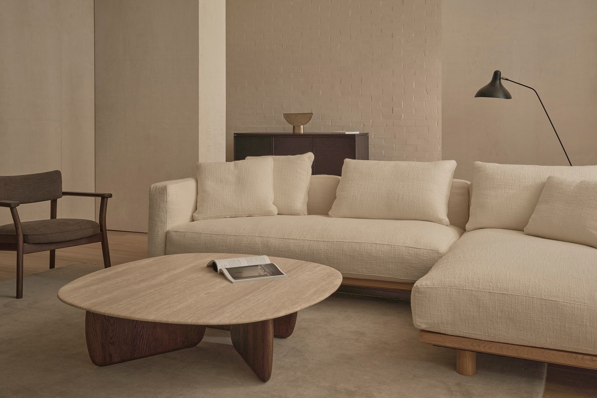 A minimalist living room features the Islet Modular Sofa | Fabric • M1 by Wooden Stories, paired with a wooden lounge chair, light wood coffee table with an open book, black wall lamp, and dark sideboard against neutral tones.