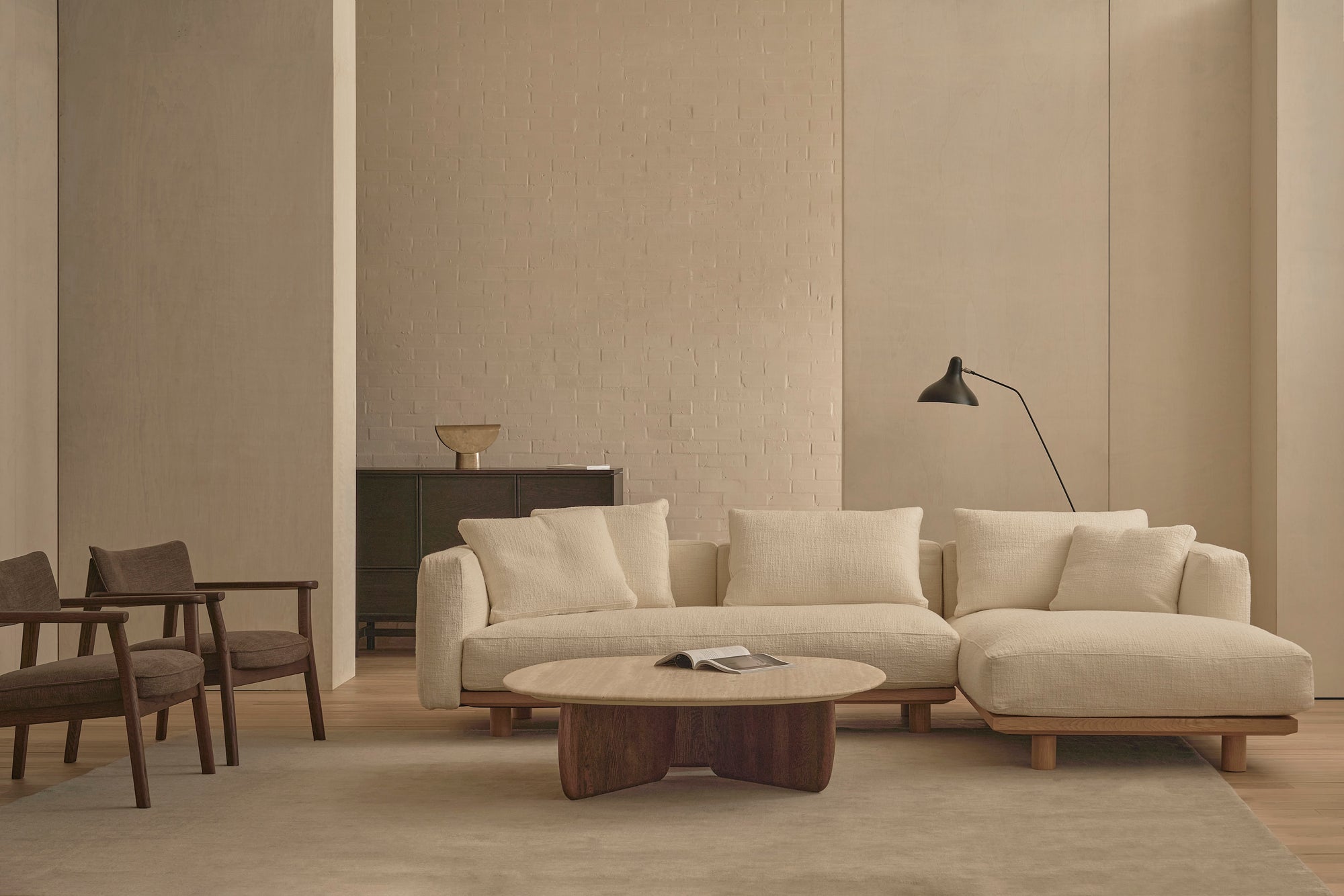 A modern living room featuring the Islet Modular Sofa | Fabric • M1 by Wooden Stories, a wooden coffee table with a book, two brown chairs, a black floor lamp, a dark sideboard, and neutral-toned walls and rug.