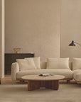 A modern living room featuring the Islet Modular Sofa | Fabric • M1 by Wooden Stories, a wooden coffee table with a book, two brown chairs, a black floor lamp, a dark sideboard, and neutral-toned walls and rug.