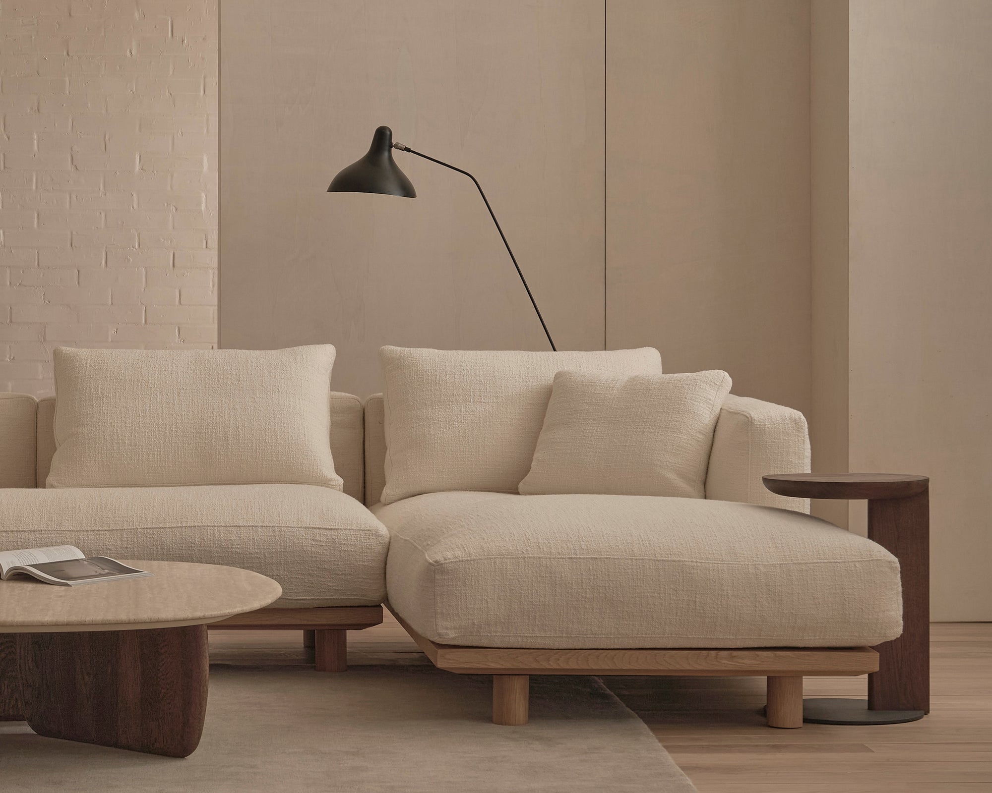 A modern living room featuring Wooden Stories' Islet Modular Sofa | Fabric • M1 in light beige, matching cushions, a round wooden coffee table with an open book, a dark side table, a black wall-mounted lamp, neutral walls, and a light rug.