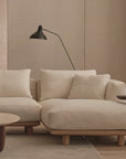 A modern living room featuring Wooden Stories' Islet Modular Sofa | Fabric • M1 in light beige, matching cushions, a round wooden coffee table with an open book, a dark side table, a black wall-mounted lamp, neutral walls, and a light rug.