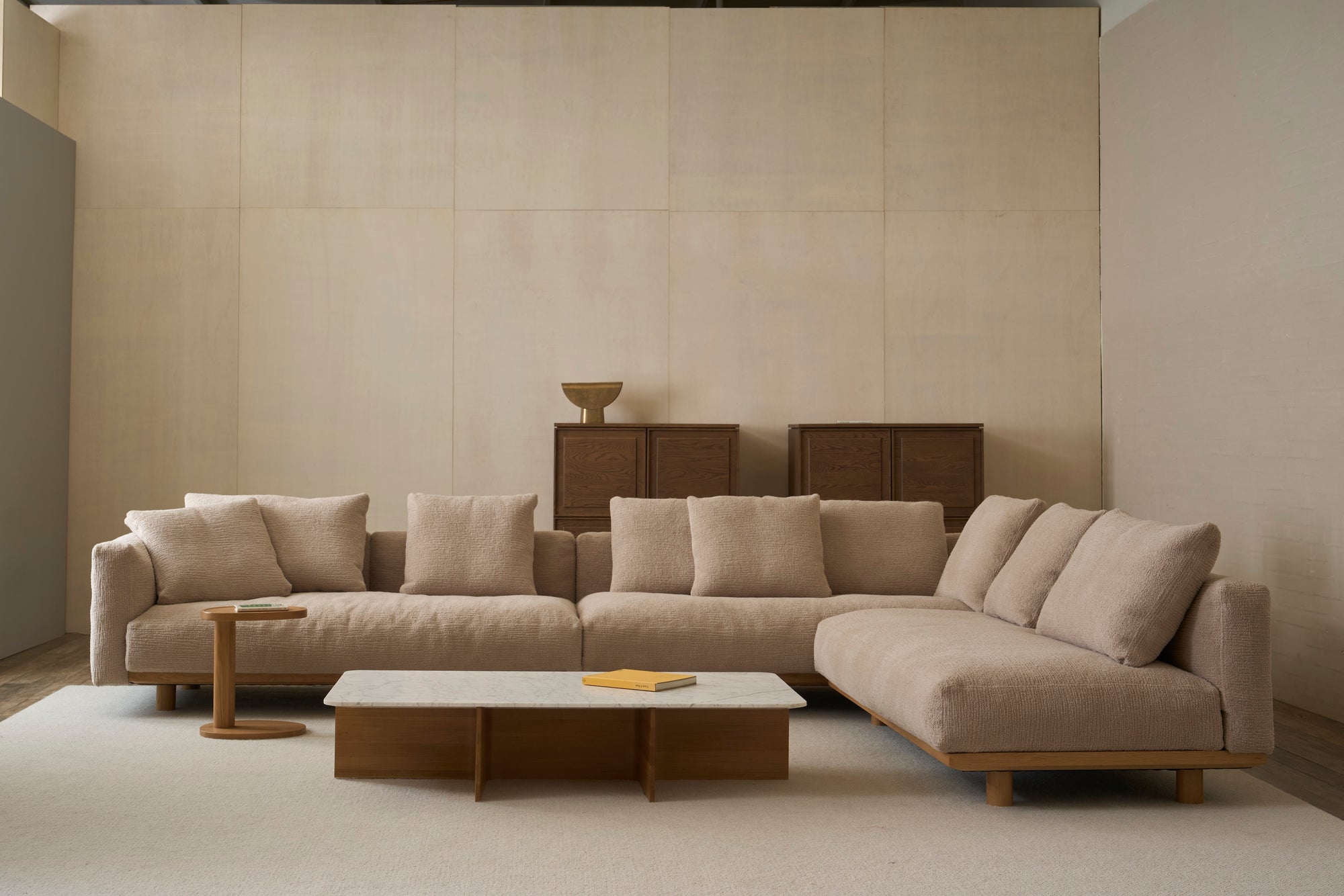 A modern living room features the Islet Modular Sofa | Fabric • M2 by Wooden Stories with plush cushions, a rectangular marble coffee table, a wooden side table, and two wooden cabinets against a neutral-toned wall.