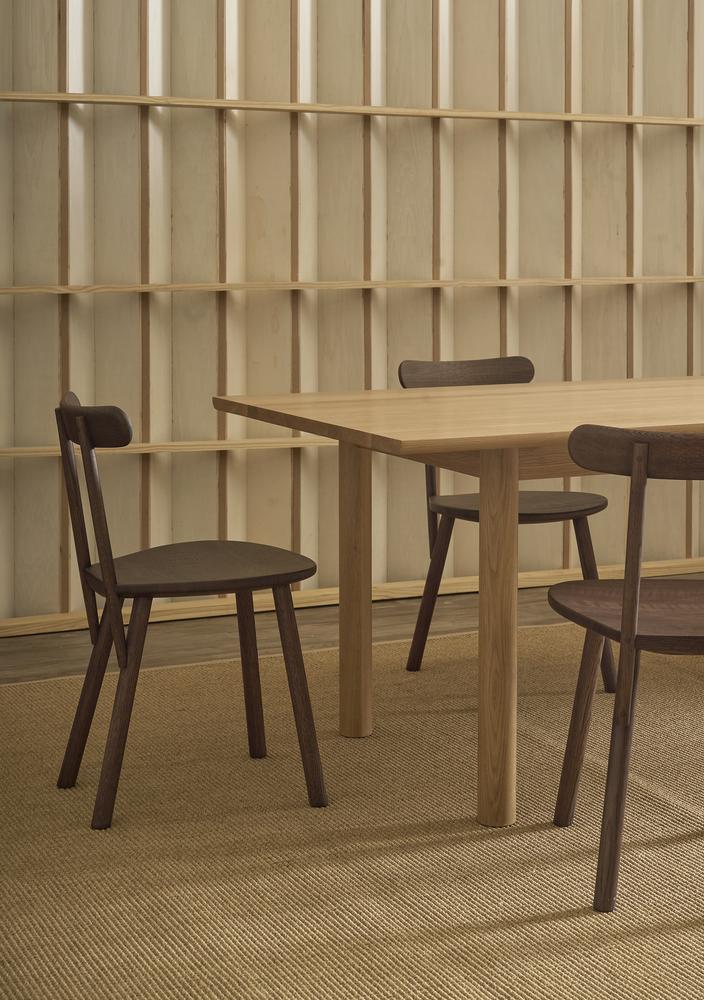 Three wooden chairs surround the Wooden Stories Itamae Dining Table in Light Oak, set in a minimalist room with light wood panel walls and a beige woven rug, reflecting modern Danish design aesthetics.