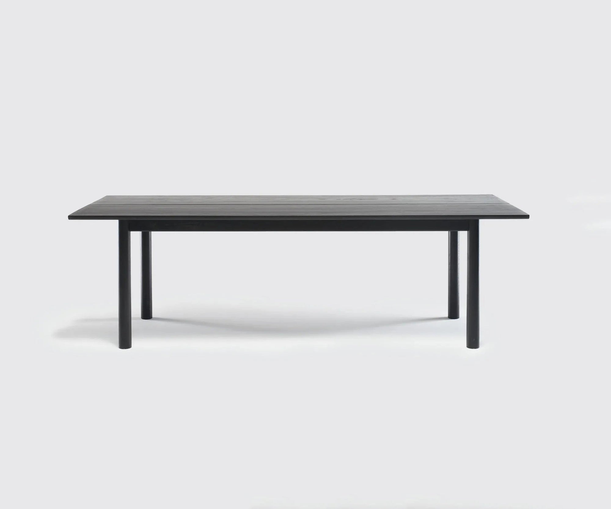 The Itamae Dining Table | Black Oak by Wooden Stories features a rectangular solid oak top and four thick cylindrical legs. Inspired by Scandinavian design, it's perfect for contract grade settings and shown against a light gray background.