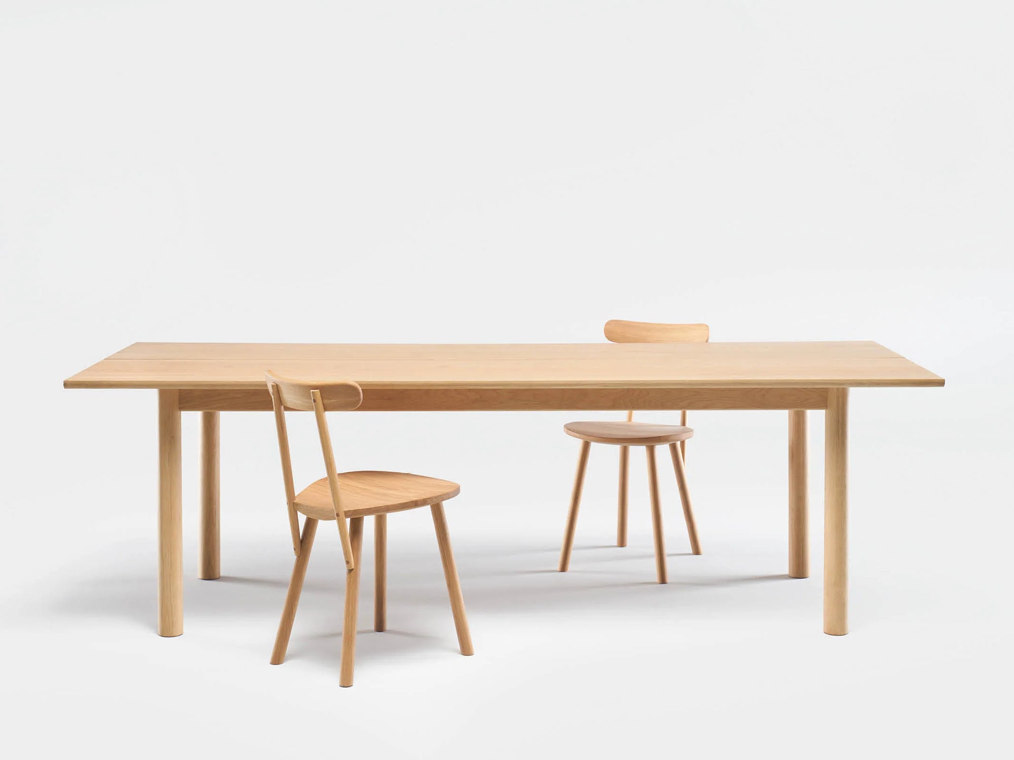 A minimalist set by Wooden Stories featuring the Itamae Dining Table in Light Oak and two matching chairs, displayed on a plain white background. One chair is tucked under the table while the other is slightly pulled out.