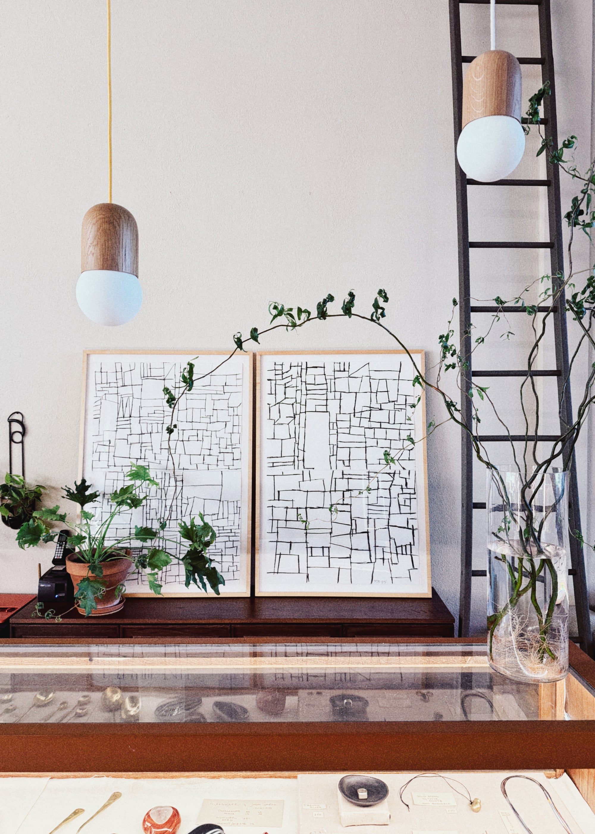 A minimalist interior featuring "Kyoto 1 and 2" charcoal on paper abstract prints by ROBERT BRADY, potted plants, a tall vase with branches, pendant lights, a glass display case with jewelry, and a black metal ladder on the right.