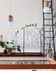 A minimalist interior featuring "Kyoto 1 and 2" charcoal on paper abstract prints by ROBERT BRADY, potted plants, a tall vase with branches, pendant lights, a glass display case with jewelry, and a black metal ladder on the right.