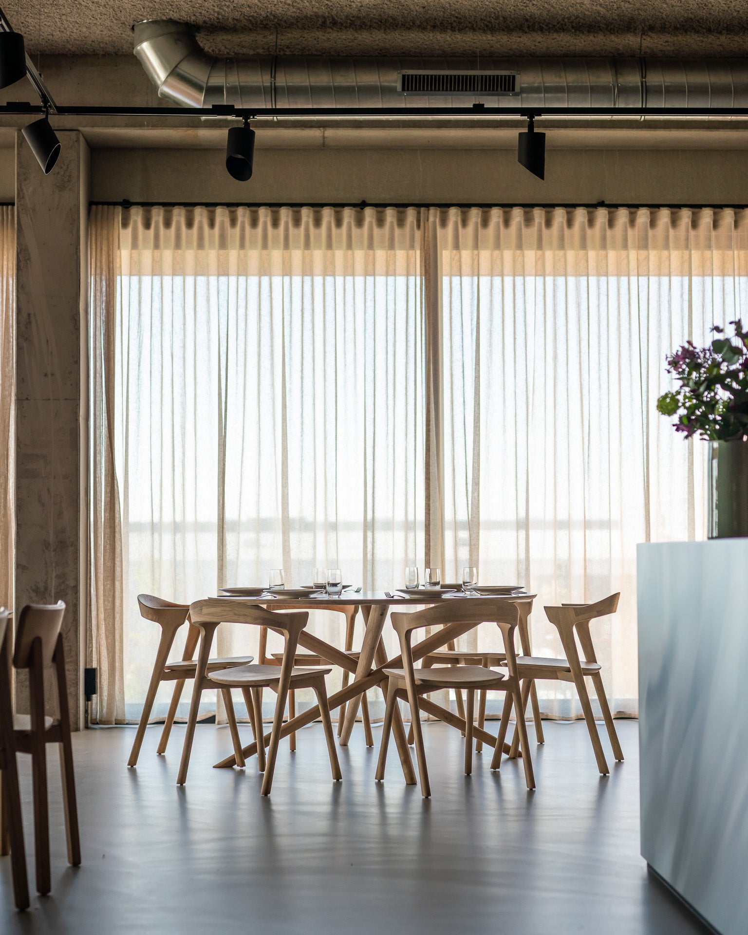 A modern dining area showcases the Ethnicraft Mikado Dining Table Round in oak, paired with wooden chairs. Sunlight streams through large windows with sheer curtains, while a vase of flowers adorns a nearby counter.