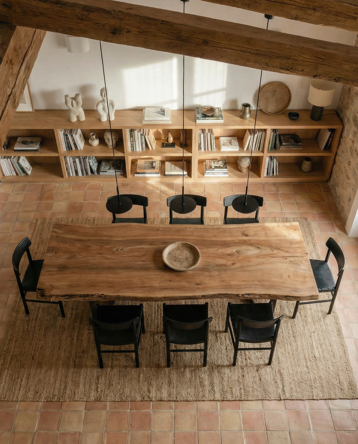 The SOBU Leviathan Live Edge Dining Table with Curved Metal Legs and seven black chairs rests on a woven rug in a rustic room with terracotta tile floors, wooden ceiling beams, shelves of books and decor, and abundant natural light.