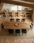 The SOBU Leviathan Live Edge Dining Table with Curved Metal Legs and seven black chairs rests on a woven rug in a rustic room with terracotta tile floors, wooden ceiling beams, shelves of books and decor, and abundant natural light.