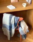A wooden chair with the Elvang Denmark Lava Throw in Cobalt Blue is placed by a wooden desk, topped with a closed notebook and a small textured ceramic cup.