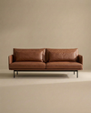 A modern Cherry Sofa | Leather by Wooden Stories, featuring rich brown leather upholstery and sleek metal legs, sits against a plain beige background. The minimalist design boasts two seat cushions and two back cushions, creating a clean, stylish look that ensures exceptional comfort—making it an ideal choice for any modern living space.
