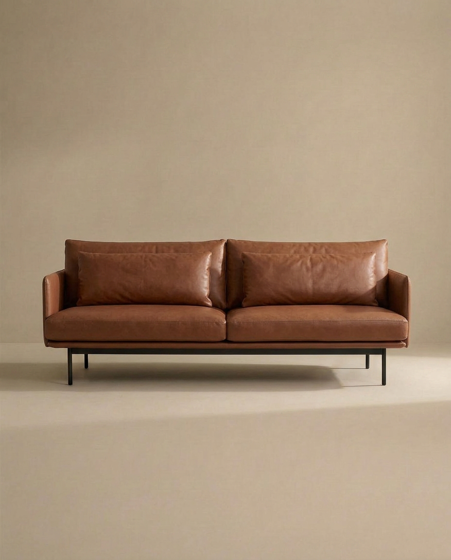 A modern Cherry Sofa | Leather by Wooden Stories, featuring rich brown leather upholstery and sleek metal legs, sits against a plain beige background. The minimalist design boasts two seat cushions and two back cushions, creating a clean, stylish look that ensures exceptional comfort—making it an ideal choice for any modern living space.