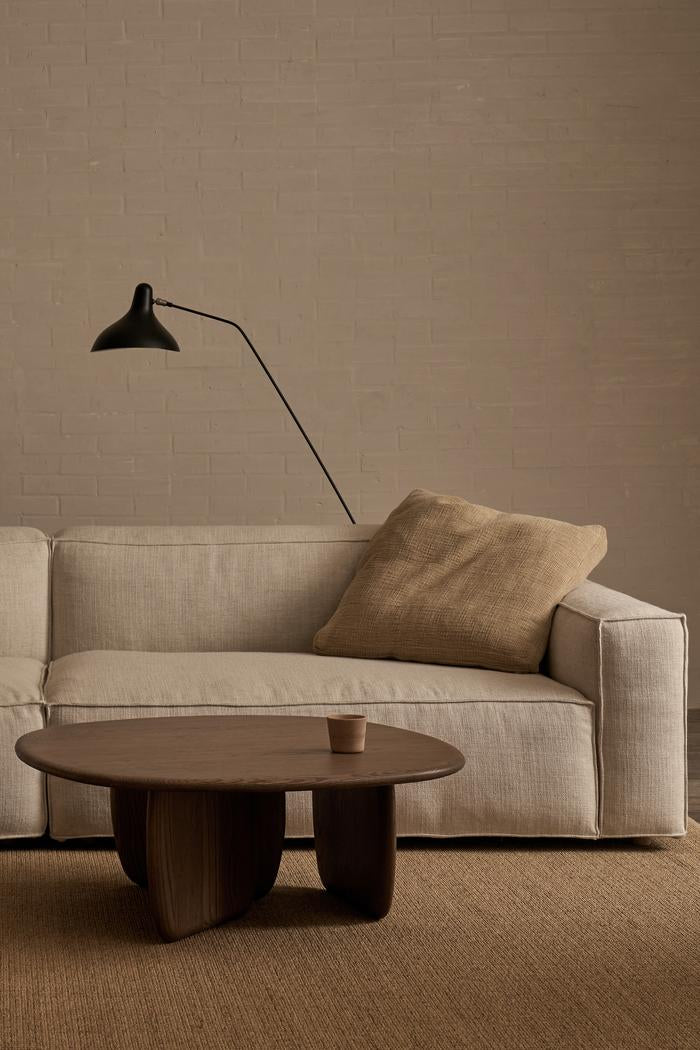 A minimalist living room with a beige sofa, textured tan cushion, modern black wall lamp, timber supports, and the Wooden Stories Eden Coffee Table in Espresso Oak with a small cup—set against a light brown brick wall and woven rug.