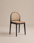 The Luk Dining Chair by Wooden Stories features a black wooden frame, curved cane weave rattan backrest, and light brown leather or faux leather seat, shown against a plain beige background.
