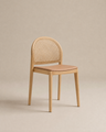 The Luk Dining Chair by Wooden Stories features a rounded cane weave rattan backrest and a light brown leather seat, shown against a plain beige background.