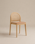 The Luk Dining Chair by Wooden Stories features a rounded cane weave rattan backrest and a light brown leather seat, shown against a plain beige background.
