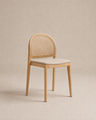 The Luk Dining Chair | Fabric Seat by Wooden Stories is a light wooden chair featuring a rounded cane weave rattan backrest and a beige upholstered seat, displayed against a neutral beige background.