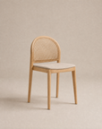 The Luk Dining Chair | Fabric Seat by Wooden Stories is a light wooden chair featuring a rounded cane weave rattan backrest and a beige upholstered seat, displayed against a neutral beige background.
