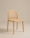 The Luk Dining Chair by Wooden Stories features a light wooden arch silhouette, rounded backrest, and woven cane seat. Set against a beige background, its minimalist design and rattan details add modern, contract-grade style.
