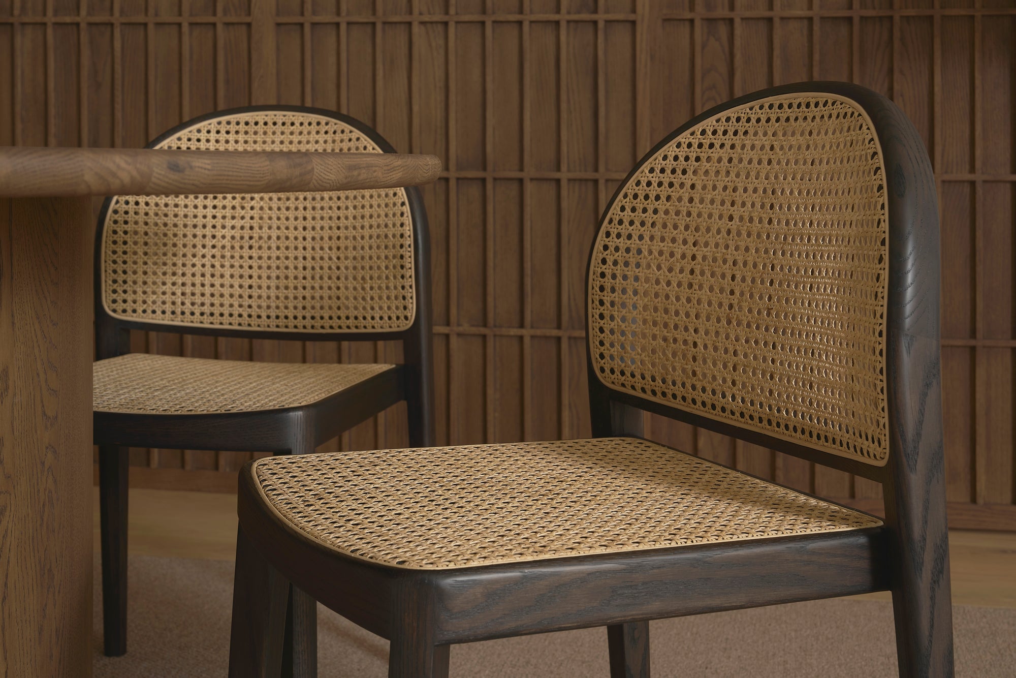 Two contract-grade Luk Dining Chairs by Wooden Stories, featuring cane-weave rattan seats and backs, are placed beside a wooden table. Vertical wood paneling in the background adds warmth to the minimalist interior.