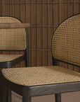 Two contract-grade Luk Dining Chairs by Wooden Stories, featuring cane-weave rattan seats and backs, are placed beside a wooden table. Vertical wood paneling in the background adds warmth to the minimalist interior.