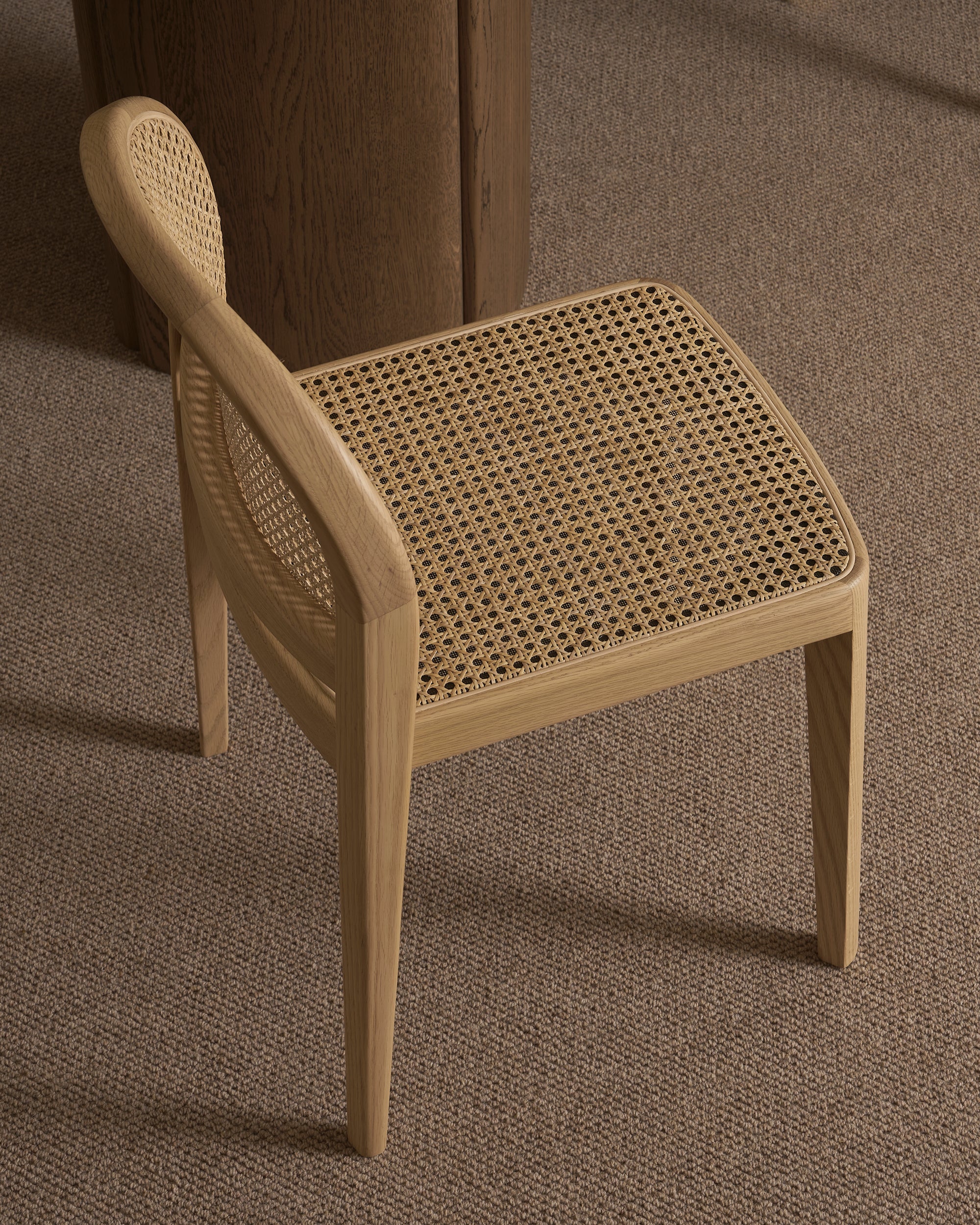 A Wooden Stories Luk Dining Chair, featuring a curved backrest and cane weave rattan seat, stands on a brown carpet beside a round wooden table.