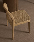 A Wooden Stories Luk Dining Chair, featuring a curved backrest and cane weave rattan seat, stands on a brown carpet beside a round wooden table.