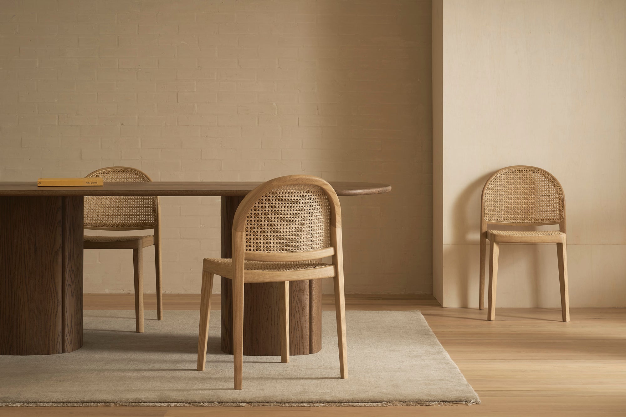 A minimalist room features a wooden table with two Wooden Stories Luk Dining Chairs and another against the wall, all in neutral tones with a light rug, soft natural lighting, and refined contract-grade furnishings.