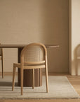 A minimalist room features a wooden table with two Wooden Stories Luk Dining Chairs and another against the wall, all in neutral tones with a light rug, soft natural lighting, and refined contract-grade furnishings.