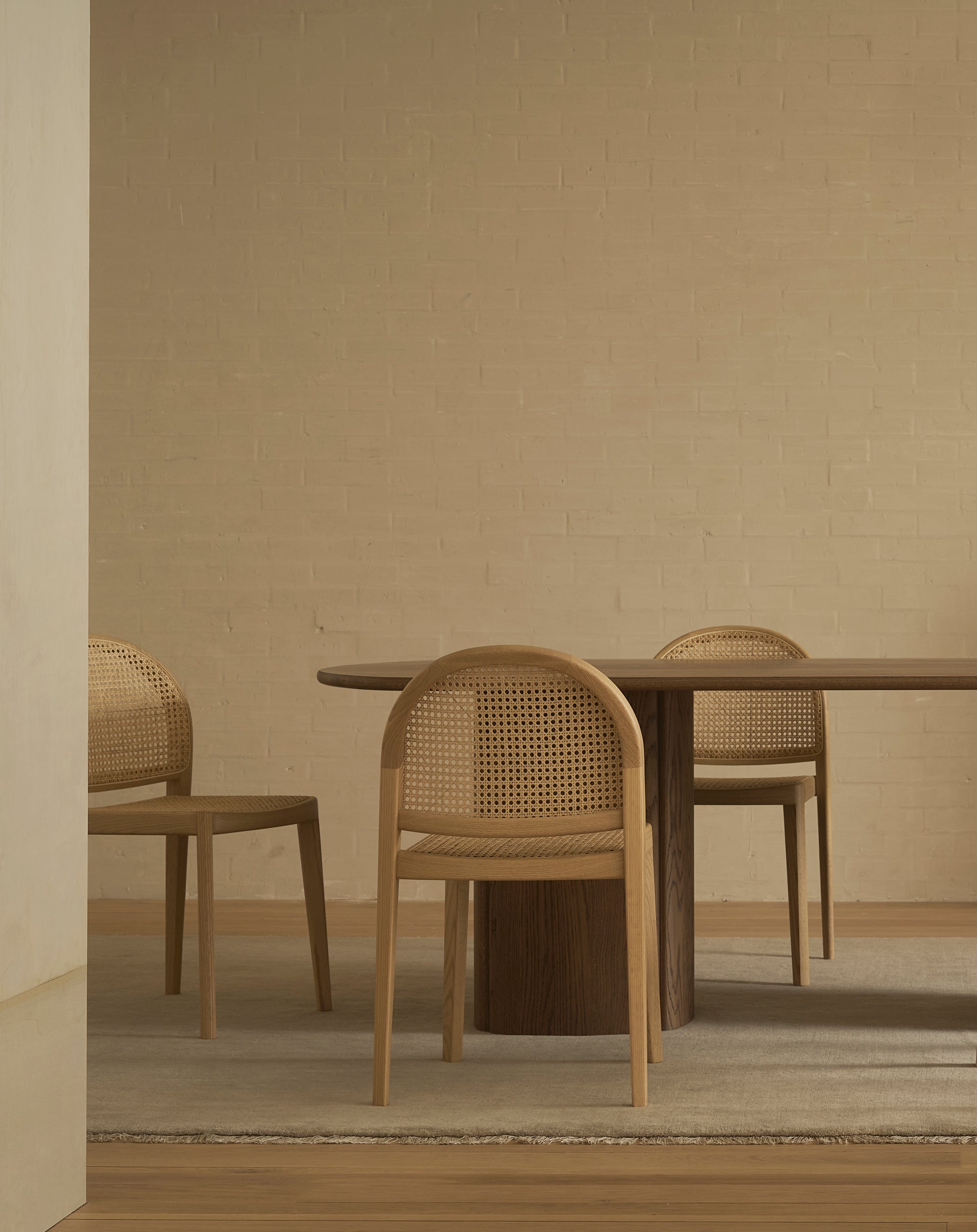 A minimalist dining room features a round wooden table and three Wooden Stories Luk Dining Chairs with cane weave rattan backs, set on a light rug near a beige brick wall. The softly lit scene is uncluttered and serene.
