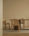 A minimalist dining room features a round wooden table and three Wooden Stories Luk Dining Chairs with cane weave rattan backs, set on a light rug near a beige brick wall. The softly lit scene is uncluttered and serene.