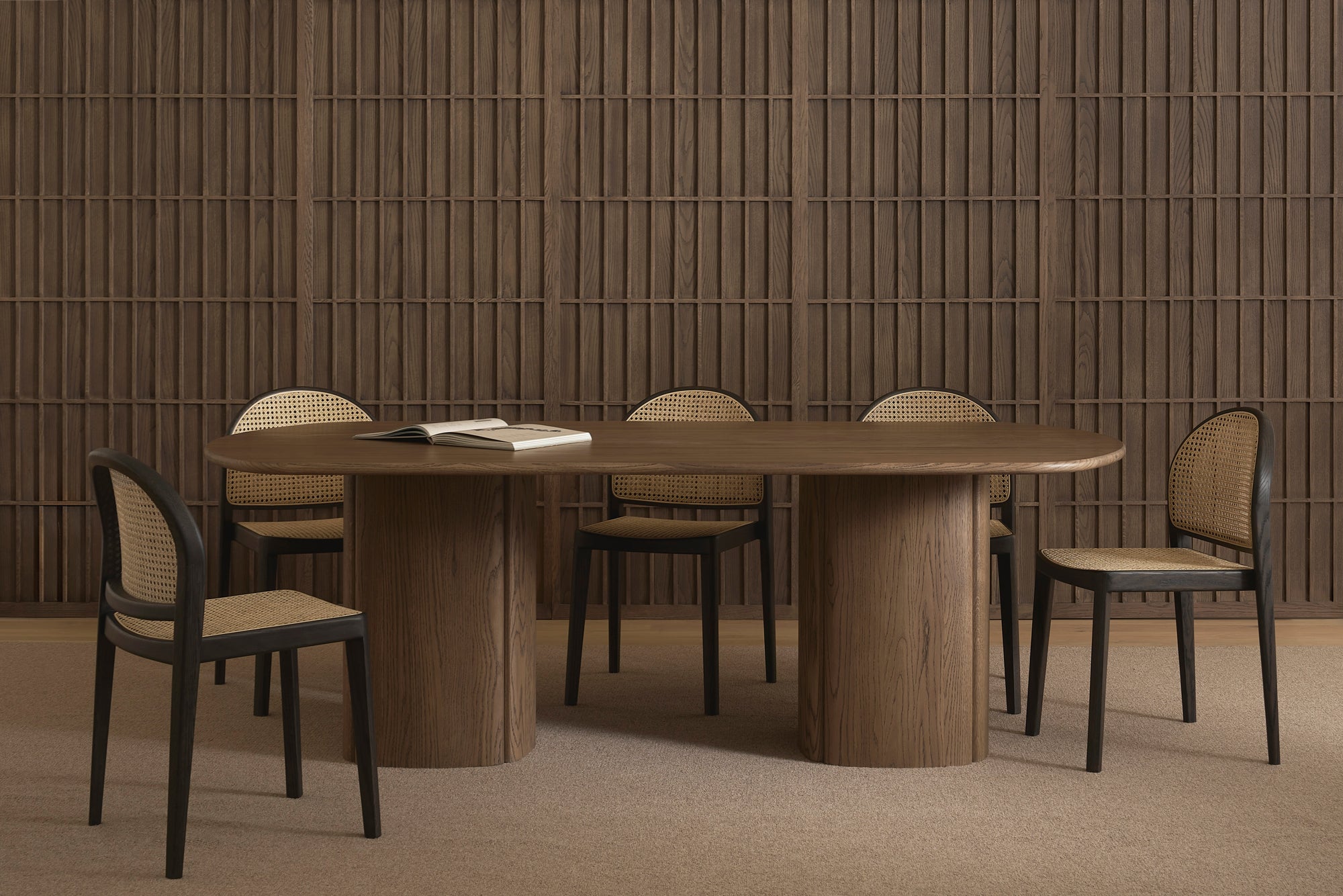 A modern dining room with a large oval wooden table and six Wooden Stories Luk Dining Chairs with cane weave rattan backs, an open book on the table, a wooden paneled wall, and minimalist decor in neutral brown tones.