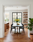 A modern dining room features a long black table, eight Wooden Stories Noki Dining Chairs in Black Oak, a vase of yellow flowers, and fruit. Large windows brighten the wood-floored space, while open shelves add warmth.