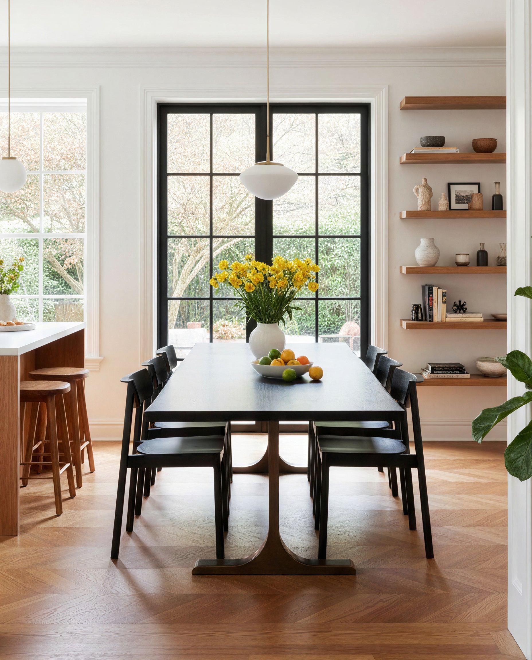 A modern dining room showcases SOBU’s Mars Live Edge Dining Table in Black Ash with Brass T Legs, paired with six black chairs, a bowl of fruit, and a yellow flower vase. Large windows bring in natural light; wooden shelves with decor line the wall.