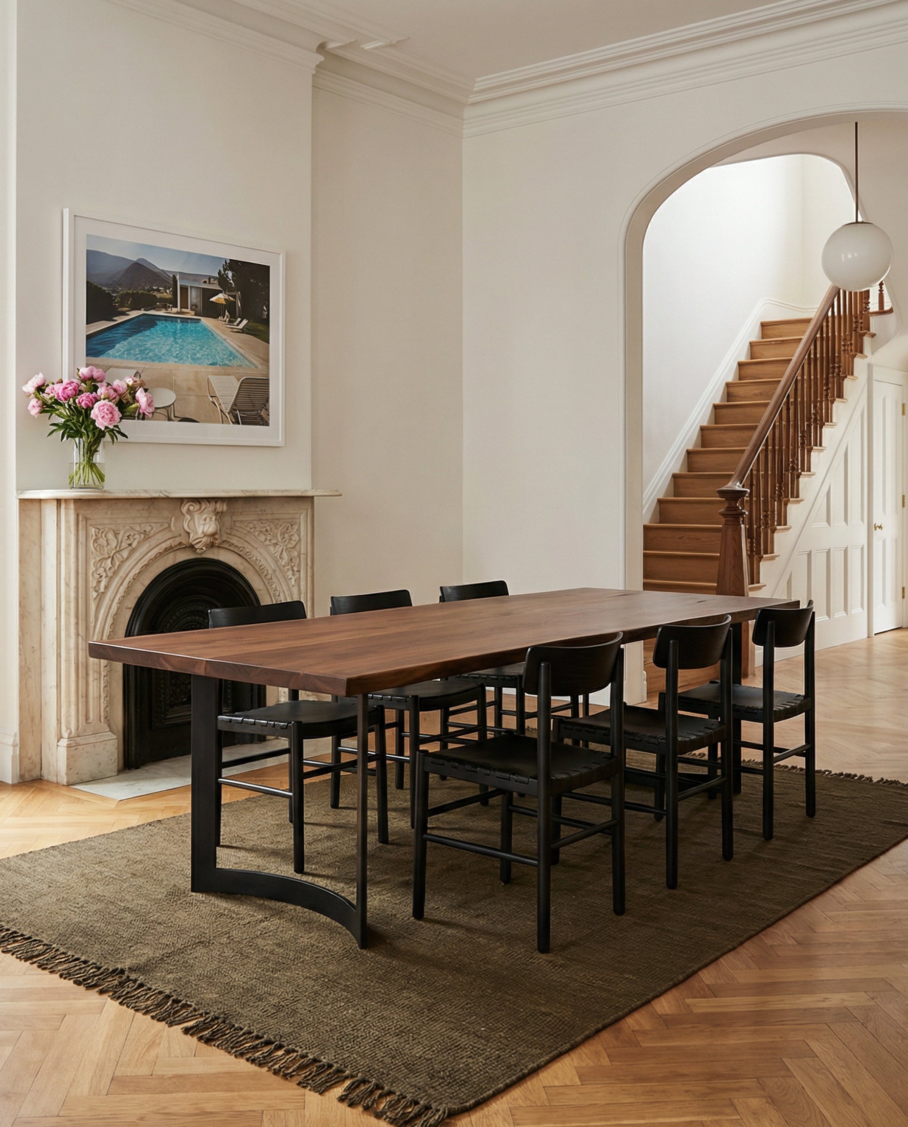 A modern dining room features the SOBU Mars Live Edge Dining Table in walnut with curved legs, eight black chairs, a brown rug, marble fireplace with a vase, framed artwork above, and a staircase with wooden banister in the background.