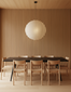A minimalist dining room features a black rectangular table, six Wooden Stories Noki Dining Chairs in Light Oak, a slatted wood wall, light wood floors and ceiling, and a large round paper lantern pendant above the table.