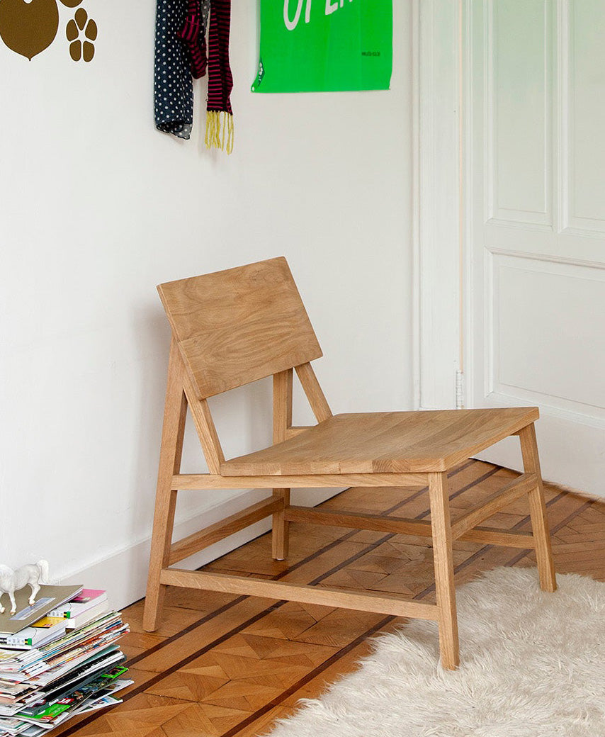 An Ethnicraft N2 Lounge Chair in solid oak with an angled backrest sits on a parquet floor by a white rug, stacked magazines nearby, and modern European design accents on the wall.