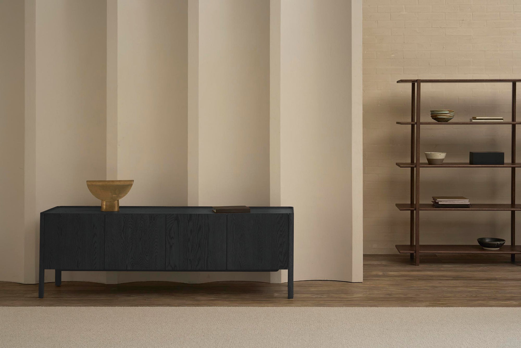 A minimalist room features the Wooden Stories Nell Sideboard in Black Oak, topped with a brass bowl and book, against a beige paneled wall. Scandinavian style shows through with wooden shelving and vessels on the warm wood floor.