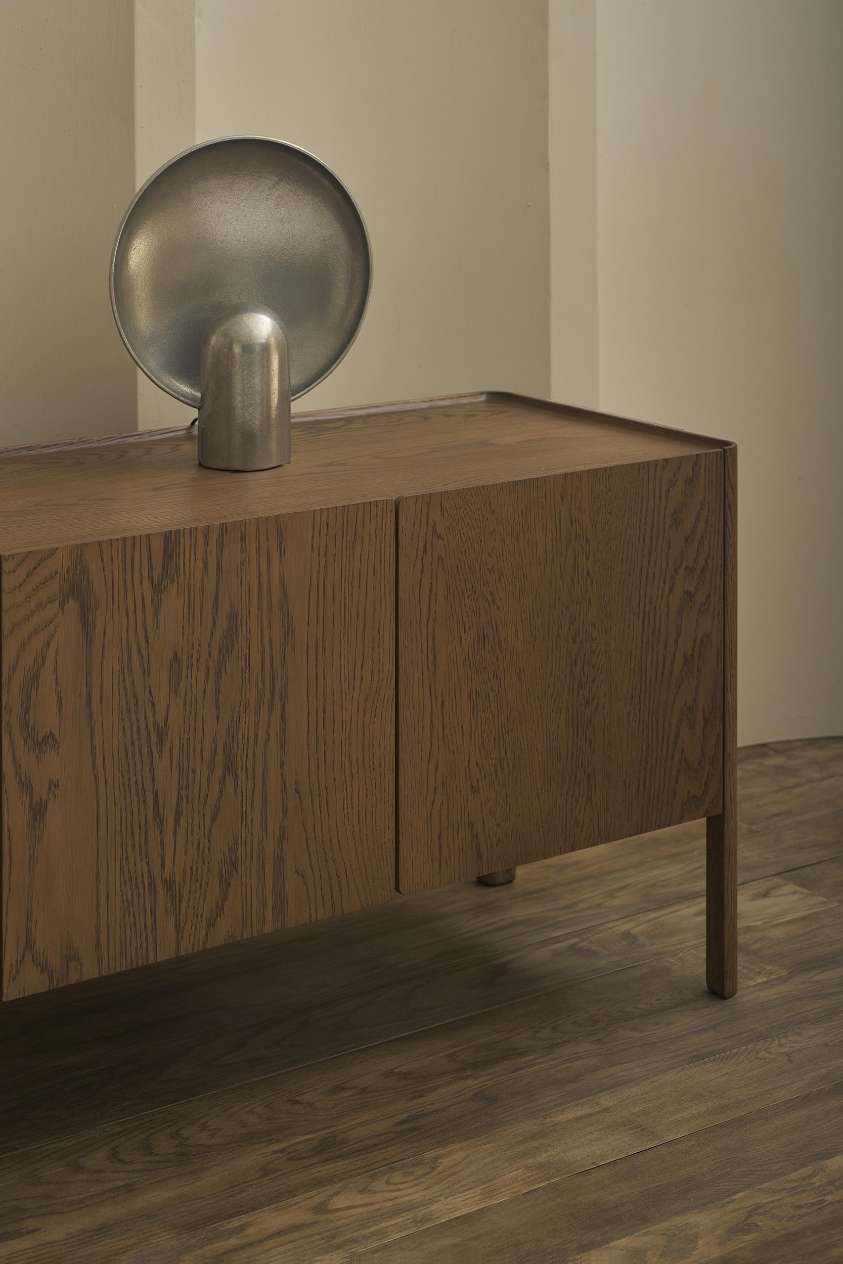 A Wooden Stories Nell Sideboard in Espresso Oak stands on a wooden floor, topped with a minimalist metallic sculpture with a rounded base and shallow oval dish, set against light walls that enhance its Scandinavian style.