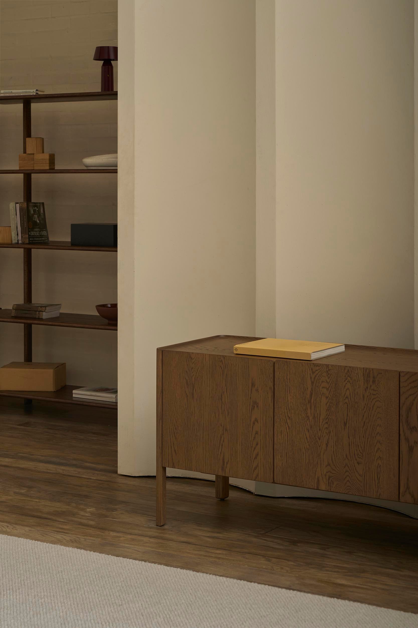 A minimalist interior features the Wooden Stories Nell Sideboard in Espresso Oak holding a closed book, a large bookshelf with books and decor, wood flooring, and a neutral rug—ideal for Scandinavian design lovers.