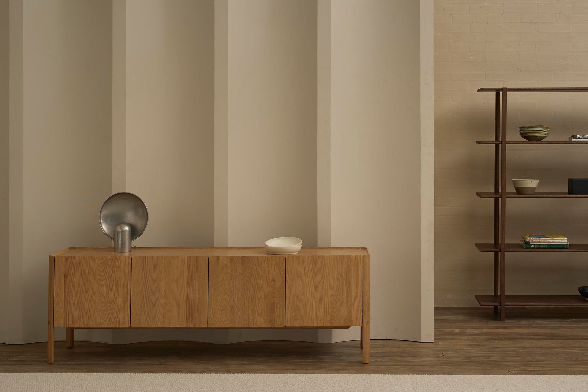 A minimalist room features the Wooden Stories Nell Sideboard | Light Oak displaying a metallic sculpture and white bowl, beside wooden shelves with bowls and books, all set against a neutral wall for a Scandinavian-inspired look.
