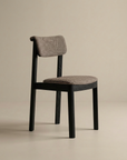 The Wooden Stories Noki Dining Chair features a contract-grade black oak frame and textured taupe fabric seat and backrest, set against a plain beige background.