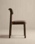 The Wooden Stories Noki Dining Chair in Espresso Oak with Fabric is shown in side view, featuring a padded, textured seat and backrest. This minimalist solid wood chair has simple lines and a natural finish, ideal for contract grade settings.