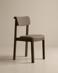 The Noki Dining Chair by Wooden Stories features an espresso oak frame with fabric-cushioned, textured seat and backrest. Its contract grade build and clean lines exemplify modern simplicity against a plain beige background.